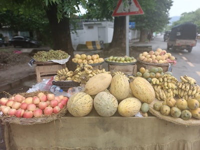 A street market stall displays a variety of fresh fruits including apples, bananas, melons, and mangoes. The fruits are arranged in baskets and on a wooden cart, with trees and a road in the background.