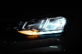 A close-up view of a car's headlight with modern design elements. The headlight is illuminated with a bright, bluish-white light and features a distinctive LED strip pattern. An amber light is also visible, adding contrast to the scene.