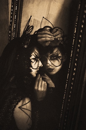A person dressed in intricate, dark lace attire gazes into a mirror, their face adorned with delicate, lace-like patterns and an ornate headpiece. The scene is dimly lit, creating a shadowy and mysterious atmosphere. The mirror reflection creates a doubling effect, adding to the surreal quality.