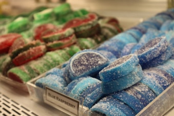 Colorful, sugar-coated gummies are displayed in a bin at a store. The image prominently features two varieties of gummies: blue raspberry-flavored in the foreground and a more assorted green and red variety in the background. Each piece is circular with a sugary texture clearly visible.