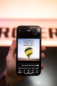 A hand holding a smartphone displaying an edited image of a hot air balloon with a motivational quote. The balloon has stripes of yellow, green, and blue, hovering above a field with mountains in the distance. Various editing tools are visible at the bottom of the phone screen.