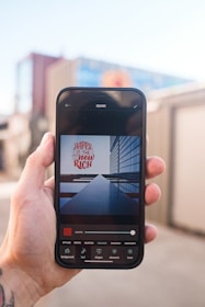 A person holds a smartphone displaying an application interface with an image editor. The edited image on the screen shows text reading 'Happy is the new rich' against a backdrop of a modern glass building. The smartphone is held outdoors, with blurred urban structures in the background.