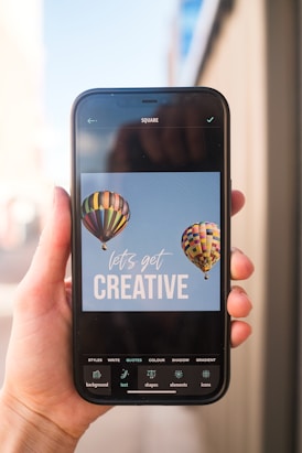 A smartphone held in a hand displays an image editing application featuring a design with two colorful hot air balloons and the phrase 'let's get creative' on a light blue background. The app interface shows various editing options at the bottom, such as styles, text, shapes, and more.