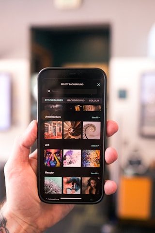 A close-up image of a smartphone being held in a hand, displaying various image categories such as Architecture, Art, and Beauty. The screen shows stock images for background selections, with thumbnails depicting different themes and art styles.