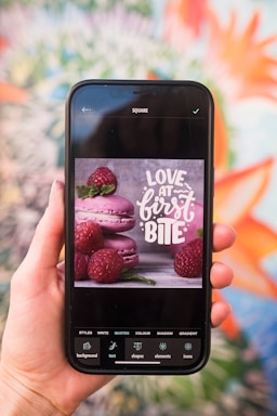 A hand holding a smartphone displaying an image of pink macarons and raspberries with text reading 'Love at First Bite.' The screen shows various editing options at the bottom, including styles, text, shapes, and elements. The background is a colorful, blurred mural with floral patterns.