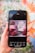 A hand holding a smartphone displaying an image of pink macarons and raspberries with text reading 'Love at First Bite.' The screen shows various editing options at the bottom, including styles, text, shapes, and elements. The background is a colorful, blurred mural with floral patterns.