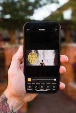 A hand holding a smartphone, displaying an application on the screen. The application showcases a rectangular image of a person standing in front of a waterfall with the text 'Energize your life' in yellow font. The background of the environment is slightly out of focus, featuring greenery and a brick wall.