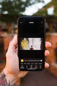 A hand holding a smartphone, displaying an application on the screen. The application showcases a rectangular image of a person standing in front of a waterfall with the text 'Energize your life' in yellow font. The background of the environment is slightly out of focus, featuring greenery and a brick wall.