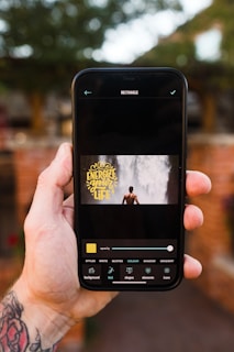 A hand holding a smartphone, displaying an application on the screen. The application showcases a rectangular image of a person standing in front of a waterfall with the text 'Energize your life' in yellow font. The background of the environment is slightly out of focus, featuring greenery and a brick wall.