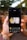 A hand holding a smartphone, displaying an application on the screen. The application showcases a rectangular image of a person standing in front of a waterfall with the text 'Energize your life' in yellow font. The background of the environment is slightly out of focus, featuring greenery and a brick wall.