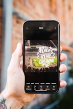 A hand holding a smartphone displaying an edited image of a stadium with a crowd, featuring the text 'Never Stop Dreaming' in large, stylized font. The phone interface shows editing options related to styles, text, and shapes.