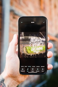 A hand holding a smartphone displaying an edited image of a stadium with a crowd, featuring the text 'Never Stop Dreaming' in large, stylized font. The phone interface shows editing options related to styles, text, and shapes.