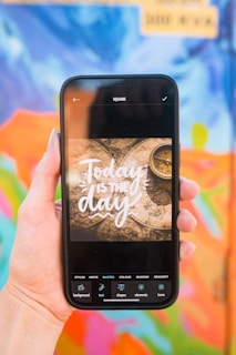 A hand holding a smartphone displaying the text 'Today is the day' over a background featuring an old map and a compass. The background behind the hand is a colorful and abstract mural with a mix of blue, orange, pink, green, and other vibrant colors.