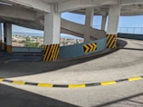 A spiraling concrete ramp inside a parking structure with black and yellow safety markings on the corners. The scene includes a view of a cityscape and blue sky in the background through open areas between the ramp's pillars.