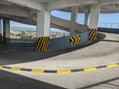 A spiraling concrete ramp inside a parking structure with black and yellow safety markings on the corners. The scene includes a view of a cityscape and blue sky in the background through open areas between the ramp's pillars.