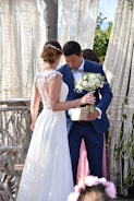 A bride in a white lace wedding gown is standing beside a groom in a blue suit holding a bouquet of white flowers and a small wooden box. They are positioned near a rustic, wooden backdrop with macramé drapes and some greenery.