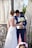 A bride in a white lace wedding gown is standing beside a groom in a blue suit holding a bouquet of white flowers and a small wooden box. They are positioned near a rustic, wooden backdrop with macramé drapes and some greenery.