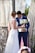 A bride in a white lace wedding gown is standing beside a groom in a blue suit holding a bouquet of white flowers and a small wooden box. They are positioned near a rustic, wooden backdrop with macramé drapes and some greenery.
