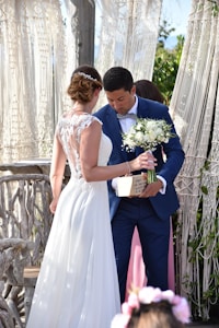 A bride in a white lace wedding gown is standing beside a groom in a blue suit holding a bouquet of white flowers and a small wooden box. They are positioned near a rustic, wooden backdrop with macramé drapes and some greenery.