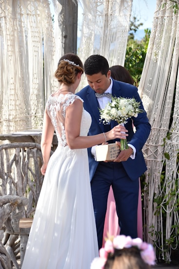 A bride in a white lace wedding gown is standing beside a groom in a blue suit holding a bouquet of white flowers and a small wooden box. They are positioned near a rustic, wooden backdrop with macram&eacute; drapes and some greenery.