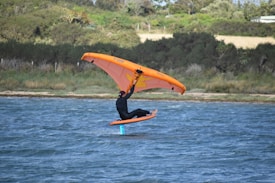 A person is engaged in wing foiling on a body of water, holding an orange wing and riding a hydrofoil board. The background features a shoreline with dense green vegetation.