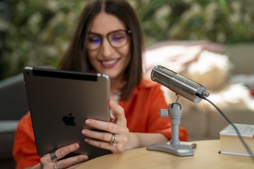 A person with glasses and a broad smile is seated at a table holding an Apple iPad. Next to them is a silver microphone on a stand, and a book on the table. The background features a blurred greenery setting, contributing to a relaxed and inviting atmosphere.