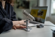 A close-up view shows a person seated at a desk with a professional microphone on a stand. The individual's hands rest on the desk near a laptop, which is open and displaying a blurred screen. The setting appears to be an indoor workspace with muted lighting.