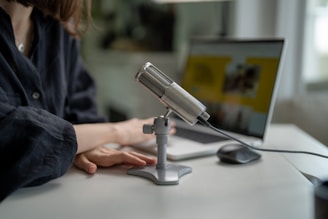 A close-up view shows a person seated at a desk with a professional microphone on a stand. The individual's hands rest on the desk near a laptop, which is open and displaying a blurred screen. The setting appears to be an indoor workspace with muted lighting.