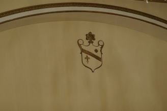 An ornate shield with a fleur-de-lis symbol beneath a decorative arch. The shield is adorned with gold and brown stripes, featuring a floral motif at the top.