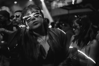 A black and white scene at a lively party or nightclub. Several people are gathered together, wearing stylish sunglasses. The lighting is dim with some bright reflections on their glasses. The focus is on a woman in the foreground, exuding a sense of confidence and enjoyment.