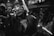 A black and white scene at a lively party or nightclub. Several people are gathered together, wearing stylish sunglasses. The lighting is dim with some bright reflections on their glasses. The focus is on a woman in the foreground, exuding a sense of confidence and enjoyment.