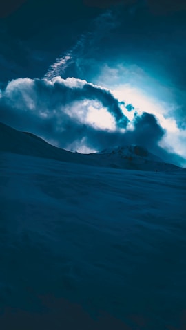 A dramatic landscape features snow-covered terrain under a sky filled with thick, swirling clouds. The lighting casts a mysterious glow, enhancing the contrast between the dark landscape and the illuminated clouds.