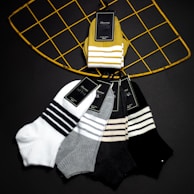 A selection of striped socks is arranged on a black background. The socks are in different colors, with labels attached, hanging from a geometric, wire grid structure painted in yellow. The colors of the socks include white, gray, mustard, and black, each having a pattern of horizontal white or black stripes.