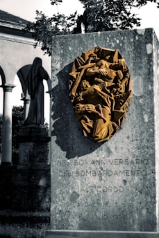 A stone monument displays a bronze relief sculpture. The relief features an abstract, dynamic composition, possibly depicting human figures entangled in turmoil. The monument's surface is inscribed with text commemorating the 50th anniversary of a bombing event on July 6, 1994. In the background, there is a statue of a cloaked figure and part of a historical building with arches and columns, with trees partially visible.