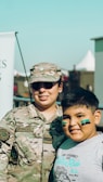A person in military camouflage uniform stands beside a smiling child with green face paint. The adult is wearing sunglasses and a cap, and the scene is outdoor with a clear sky in the background.