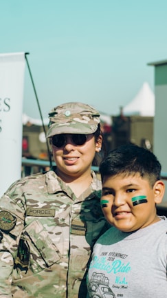 A person in military camouflage uniform stands beside a smiling child with green face paint. The adult is wearing sunglasses and a cap, and the scene is outdoor with a clear sky in the background.
