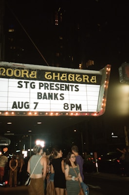 A vibrant scene at night with a group of people gathered in front of a theater marquee that reads 'STG Presents Banks Aug 7 8PM'. The street is dimly lit, with the theater sign illuminated, creating a lively urban atmosphere.