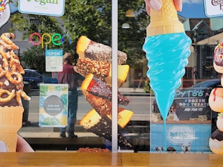 A storefront window displays large images of various ice cream treats, including a chocolate-dipped cone with pretzels, chocolate-covered ice cream bars with nuts, and a bright blue ice cream cone. Neon 'OPEN' sign and hiring notice are visible. Some reflections of people and street scenery are seen on the glass.