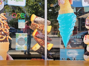 A storefront window displays large images of various ice cream treats, including a chocolate-dipped cone with pretzels, chocolate-covered ice cream bars with nuts, and a bright blue ice cream cone. Neon 'OPEN' sign and hiring notice are visible. Some reflections of people and street scenery are seen on the glass.