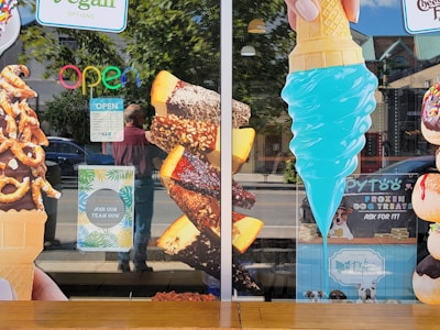 A storefront window displays large images of various ice cream treats, including a chocolate-dipped cone with pretzels, chocolate-covered ice cream bars with nuts, and a bright blue ice cream cone. Neon 'OPEN' sign and hiring notice are visible. Some reflections of people and street scenery are seen on the glass.