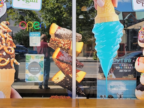 A storefront window displays large images of various ice cream treats, including a chocolate-dipped cone with pretzels, chocolate-covered ice cream bars with nuts, and a bright blue ice cream cone. Neon 'OPEN' sign and hiring notice are visible. Some reflections of people and street scenery are seen on the glass.