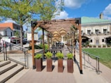 An outdoor space features a wooden pergola adorned with hanging wicker lamps and climbing plants. Below, tables and chairs offer a seating area on a patio lined with planters containing greenery. Surrounding the pergola are landscaped areas with trees and a neatly maintained lawn, bordered by an upscale building with large windows and more plants in decorative pots.
