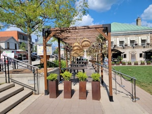 An outdoor space features a wooden pergola adorned with hanging wicker lamps and climbing plants. Below, tables and chairs offer a seating area on a patio lined with planters containing greenery. Surrounding the pergola are landscaped areas with trees and a neatly maintained lawn, bordered by an upscale building with large windows and more plants in decorative pots.