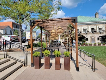 An outdoor space features a wooden pergola adorned with hanging wicker lamps and climbing plants. Below, tables and chairs offer a seating area on a patio lined with planters containing greenery. Surrounding the pergola are landscaped areas with trees and a neatly maintained lawn, bordered by an upscale building with large windows and more plants in decorative pots.