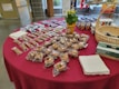 A table is covered with a red cloth and holds a variety of packaged baked goods. There are several rows of individually wrapped muffins, granola bars, and small cakes. A basket containing disposable gloves, a potted plant with yellow flowers, and boxes of juice are also visible. Stacks of napkins are neatly placed on the table.