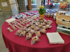 A table is covered with a red cloth and holds a variety of packaged baked goods. There are several rows of individually wrapped muffins, granola bars, and small cakes. A basket containing disposable gloves, a potted plant with yellow flowers, and boxes of juice are also visible. Stacks of napkins are neatly placed on the table.
