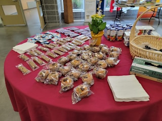 A table is covered with a red cloth and holds a variety of packaged baked goods. There are several rows of individually wrapped muffins, granola bars, and small cakes. A basket containing disposable gloves, a potted plant with yellow flowers, and boxes of juice are also visible. Stacks of napkins are neatly placed on the table.