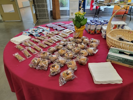 A table is covered with a red cloth and holds a variety of packaged baked goods. There are several rows of individually wrapped muffins, granola bars, and small cakes. A basket containing disposable gloves, a potted plant with yellow flowers, and boxes of juice are also visible. Stacks of napkins are neatly placed on the table.
