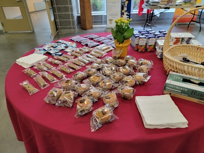A table is covered with a red cloth and holds a variety of packaged baked goods. There are several rows of individually wrapped muffins, granola bars, and small cakes. A basket containing disposable gloves, a potted plant with yellow flowers, and boxes of juice are also visible. Stacks of napkins are neatly placed on the table.