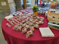 A table is covered with a red cloth and holds a variety of packaged baked goods. There are several rows of individually wrapped muffins, granola bars, and small cakes. A basket containing disposable gloves, a potted plant with yellow flowers, and boxes of juice are also visible. Stacks of napkins are neatly placed on the table.